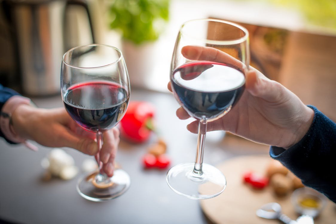 Deux verres de vin rouge trinqués par deux personnes, mains visibles, ambiance conviviale et tablette avec aliments en arrière-plan.