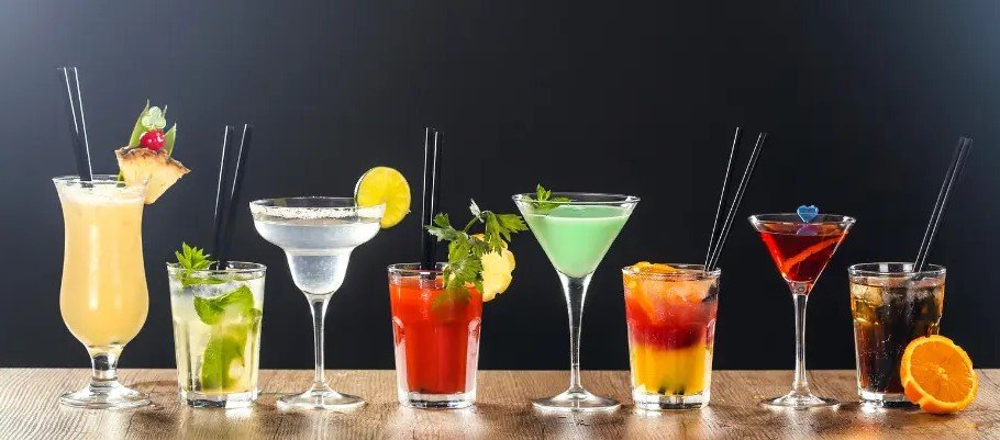 Assortiment de cocktails colorés alignés sur un plan de bois, chacun avec guirlîtes et garnitures verres variés.